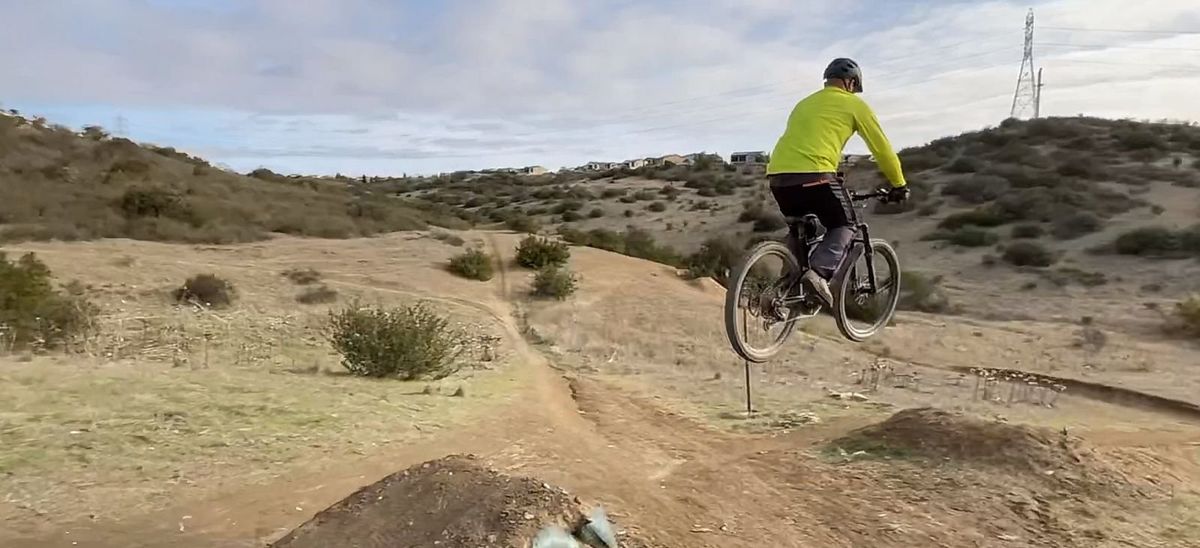 A mountain biker in a yellow jersey mid-jump on a dirt trail, with chaparral hills and houses behind